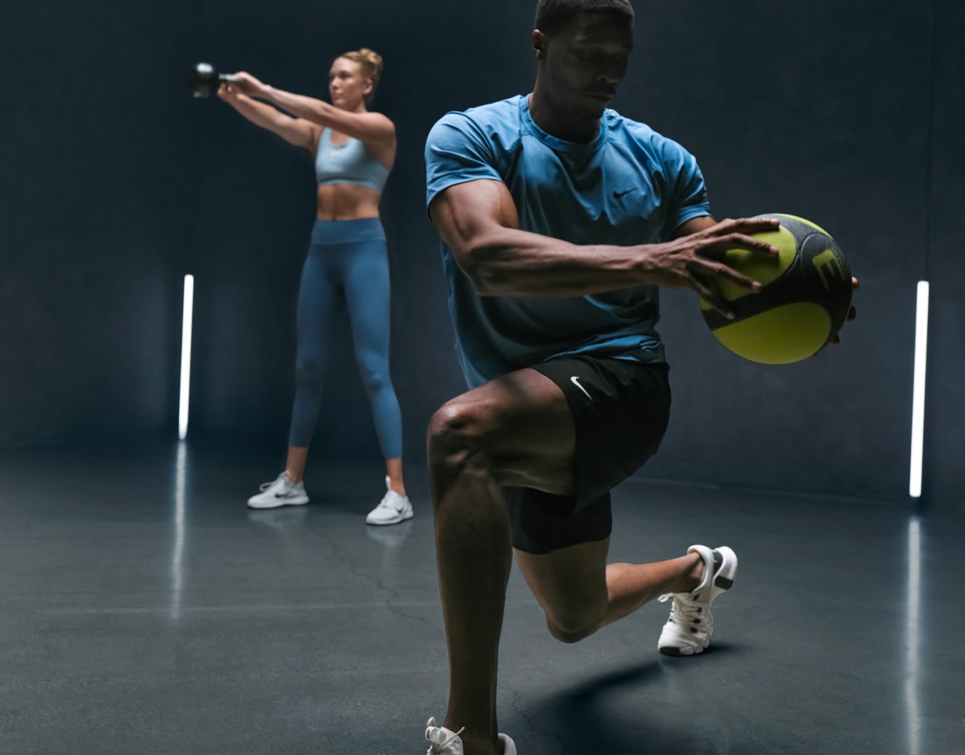 Man and woman in gym using kettle bell and weighted ball to perform athletic exercises.