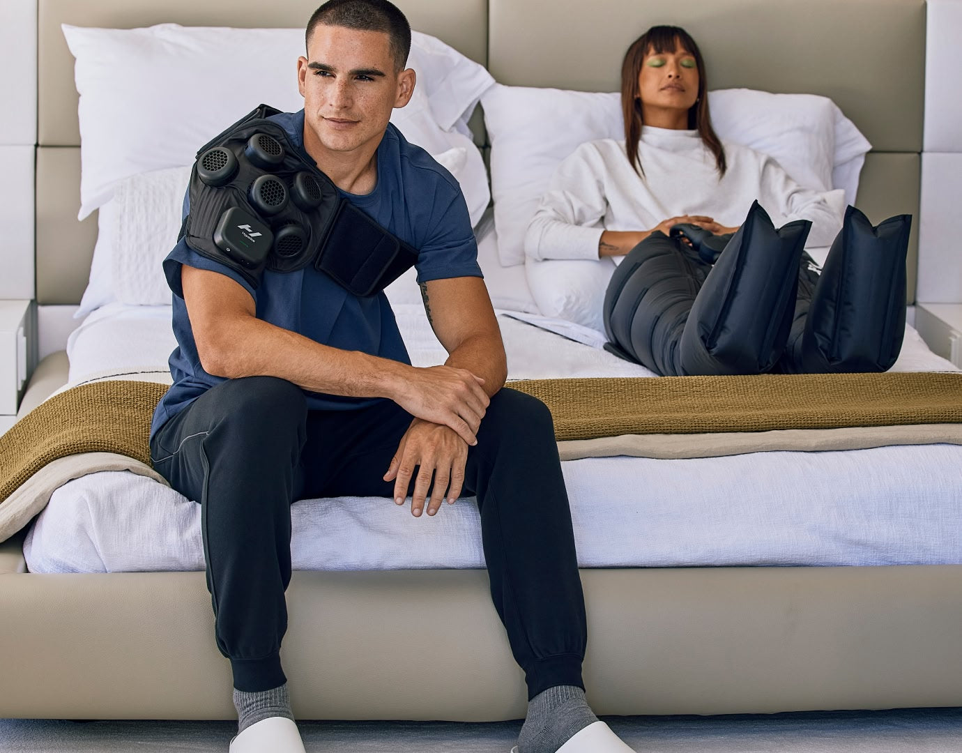Man and woman sitting on bed using Hyperice X Shoudler and Normatec 3 Legs to recover.