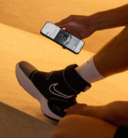 Personne portant des Hyperboot Nike x Hyperice sur un fond aux tons chauds, tenant un smartphone.