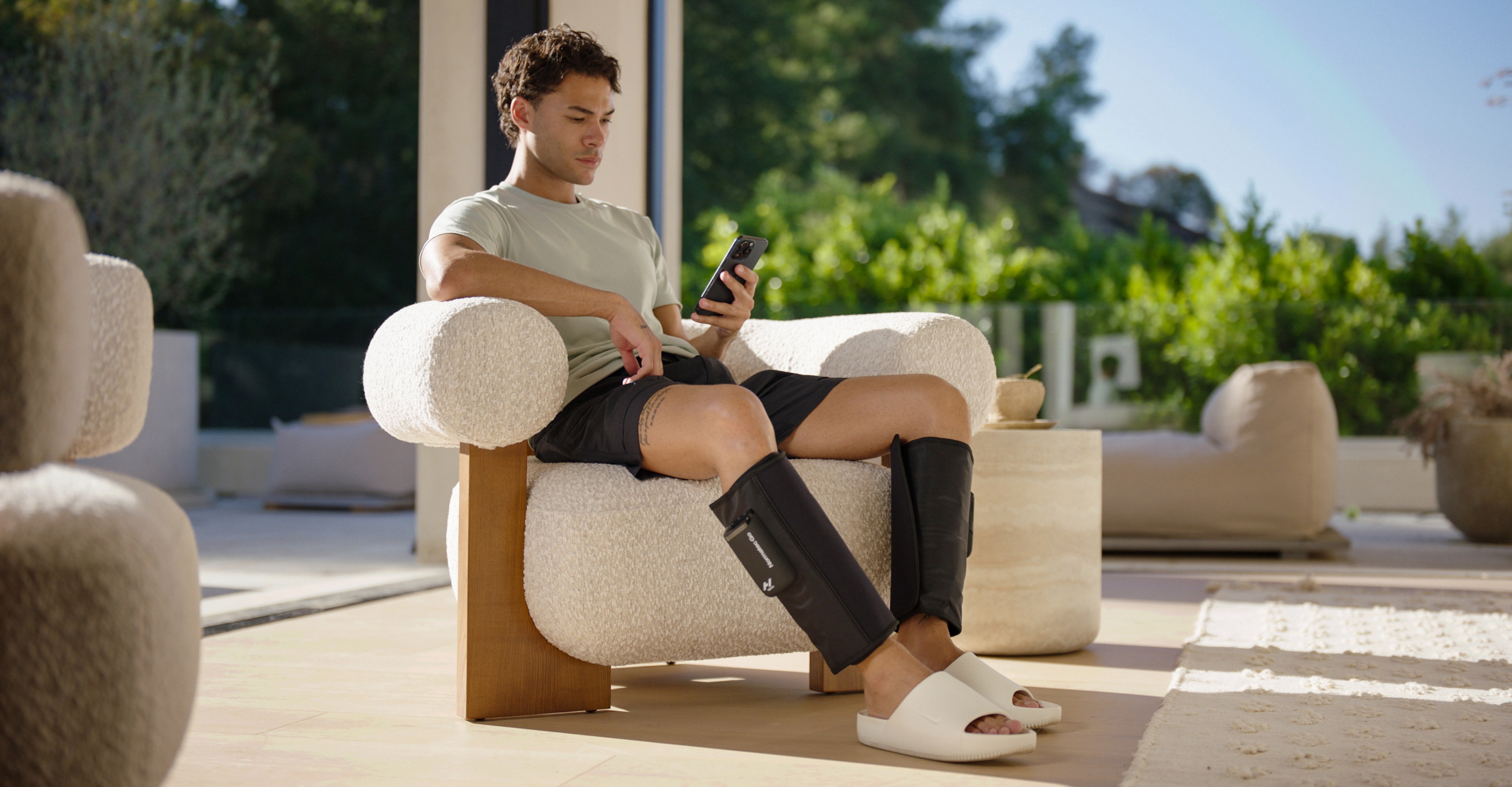 Man sitting on a couch using a smartphone wearing Normec Go compression sleeves on his calves with a green outdoor background