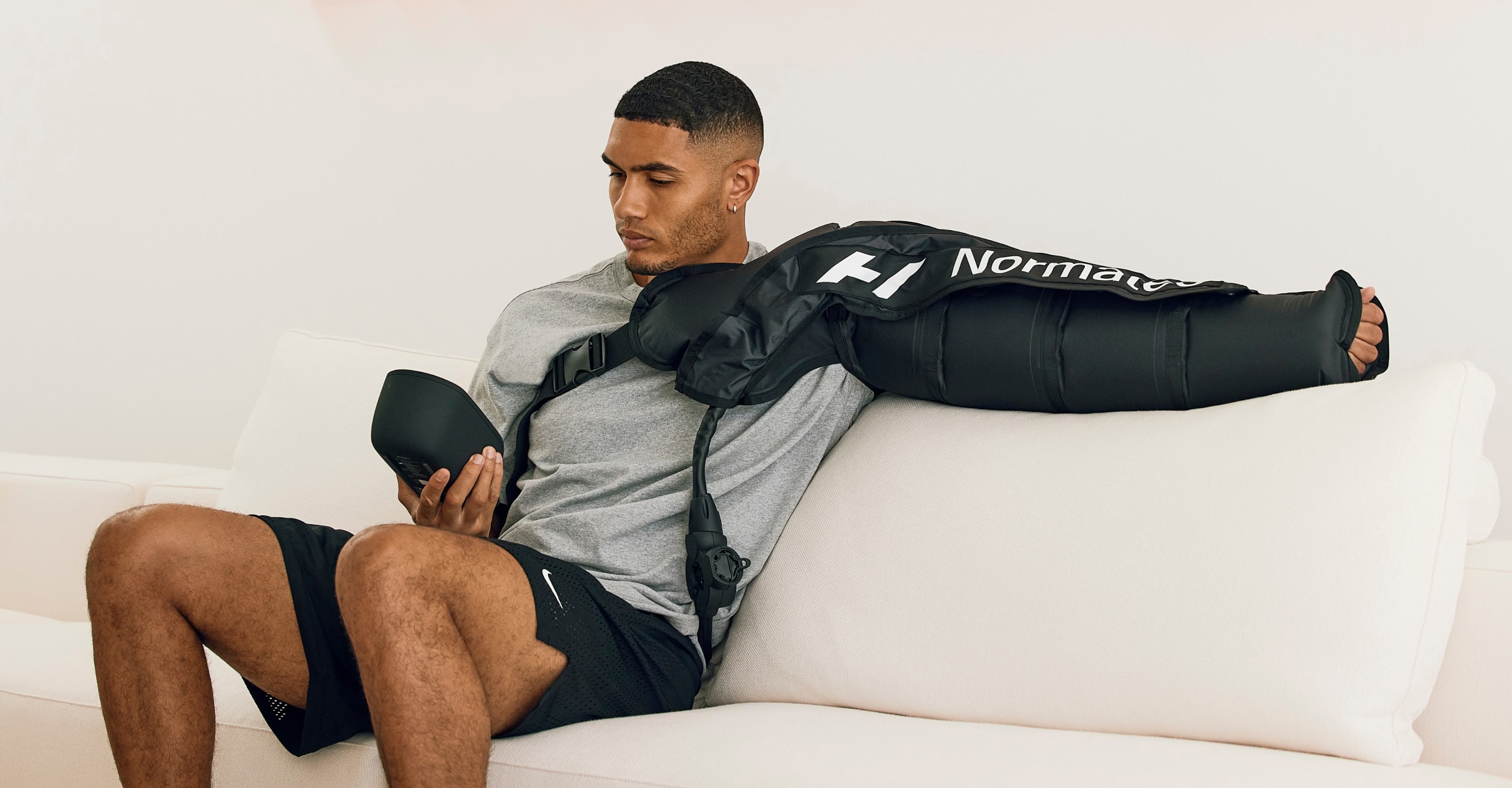 Man sitting on a couch wearing the Normatec 3 full body arm attachment, while holding the control unit.