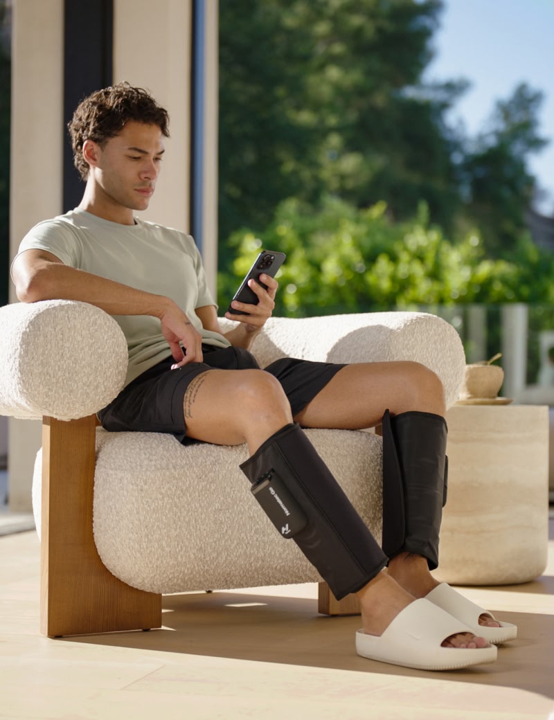 Man sitting on a chair using a smartphone wearing normatec go compression devices on his calves with a blurred outdoor background