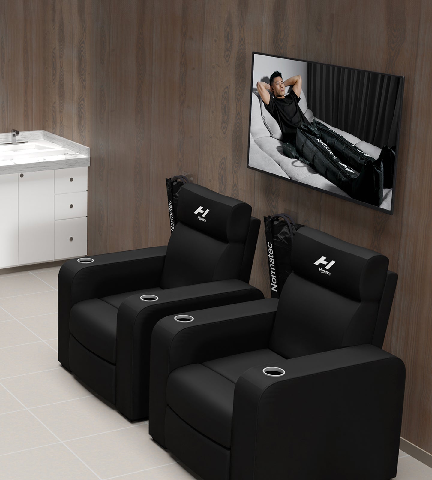 Fauteuils inclinables noirs de la marque Hyperice dans une pièce avec une télévision affichant une personne sur un canapé utilisant son appareil normatec 3.