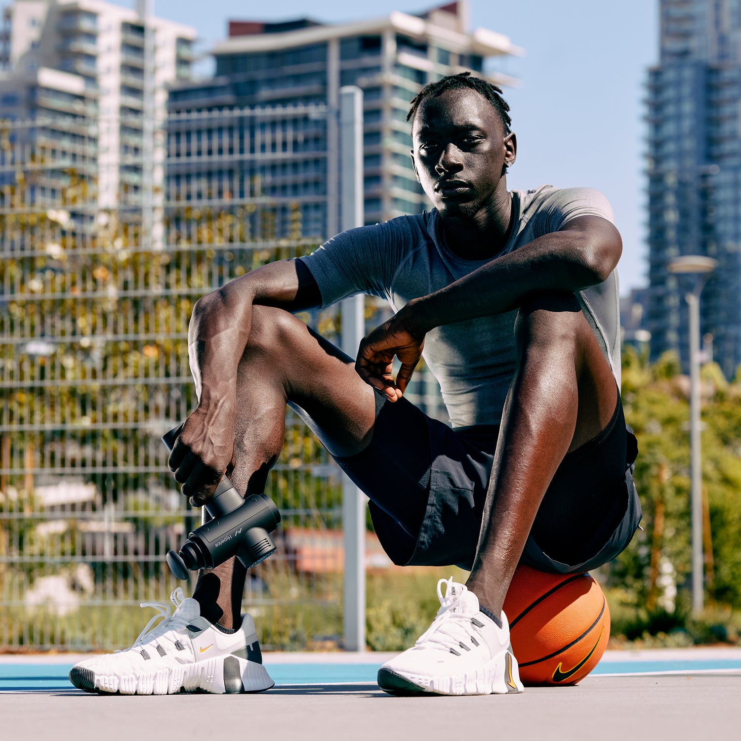 Man sitting on a basketball holding a Hypervolt with a cityscape in the background