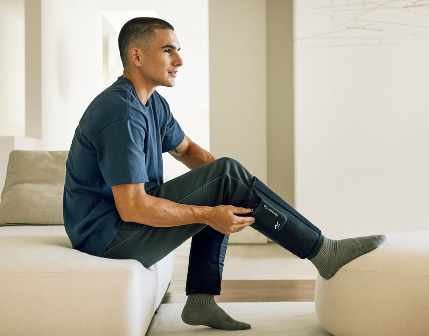 Man on couch using Normatec Go compression sleeves on his legs.