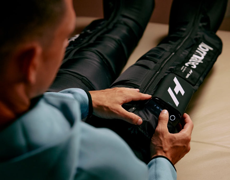 Over shoudler image of man on couch wearing normatec elite leg compression sleeves on legs.