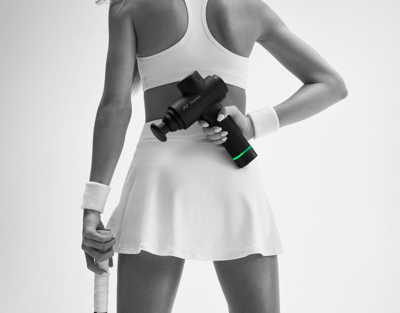 Woman in white tennis outfit holding hypervolt massage gun behind her back.