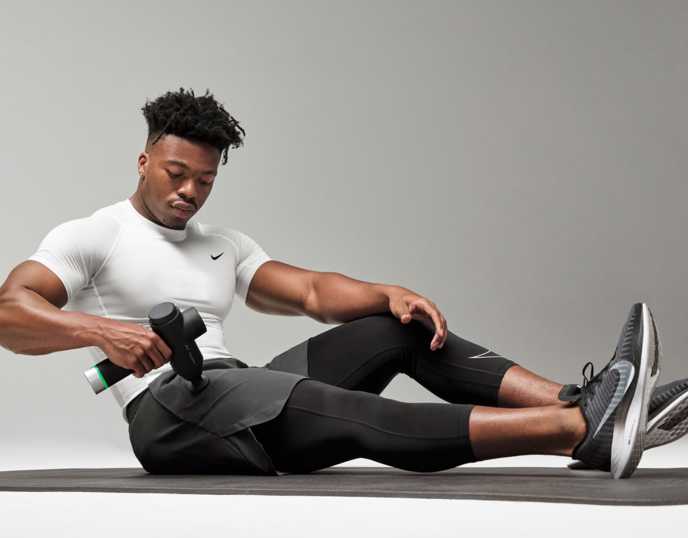 Man sitting on yoga mat using Hypervolt massage gun on right upper thigh near hip flexor.