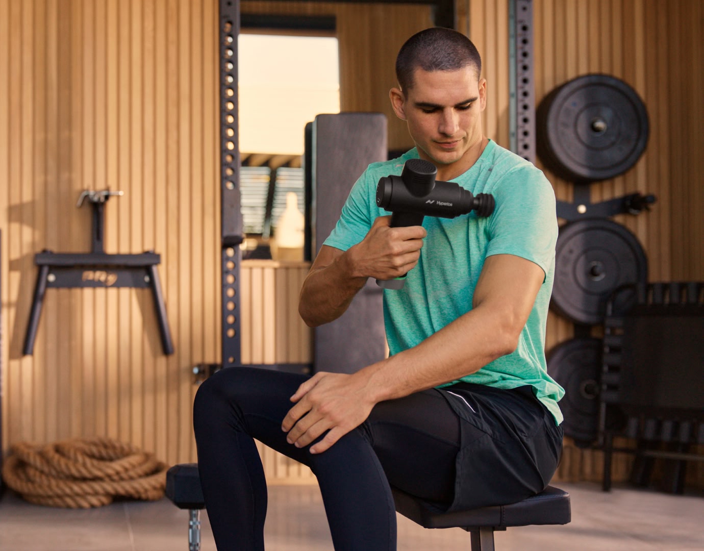 Man seated on workout bench using hypervolt massage gun on left shoulder muscle.