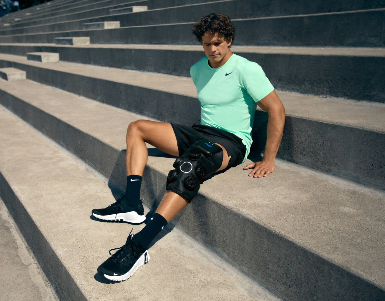Man seated on stadium steps wearing Hyperice X 2 knee on left knee. 
