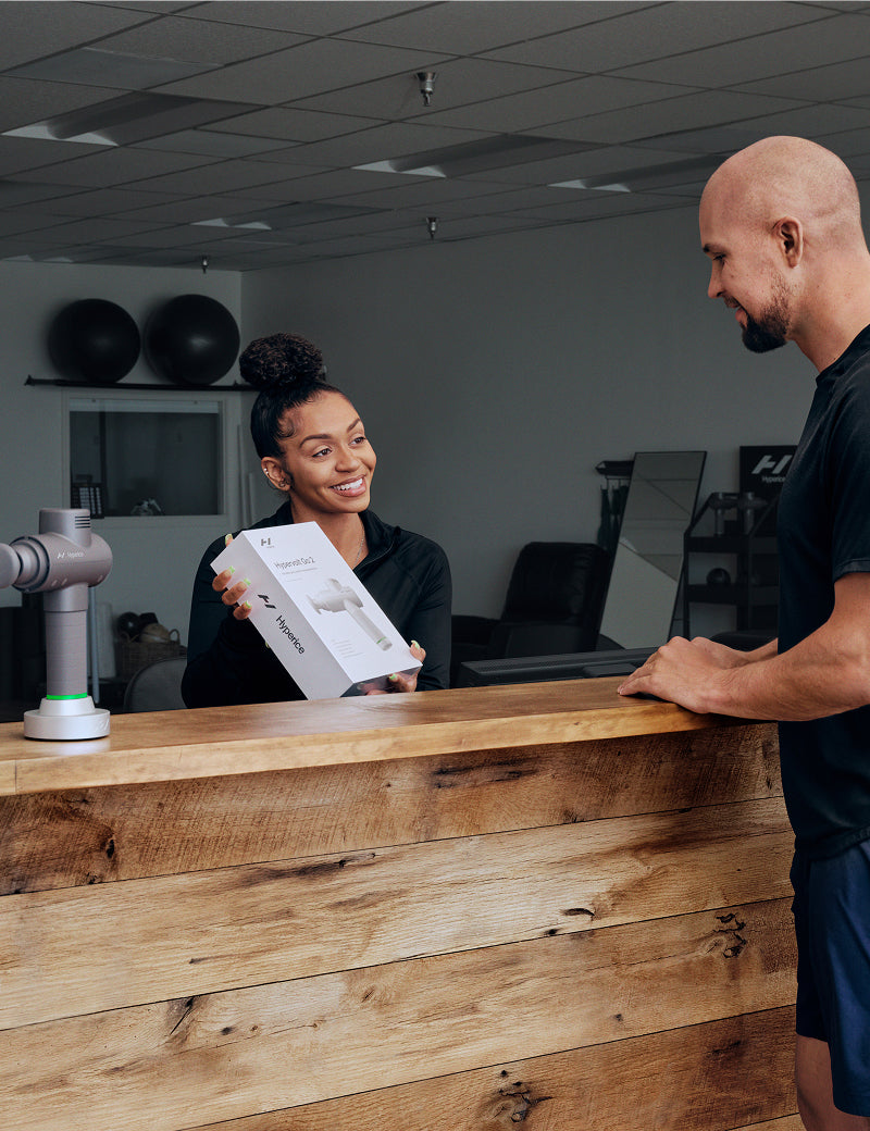 Person receiving a Hypervolt Go 2 from a receptionist at a gym reception desk.