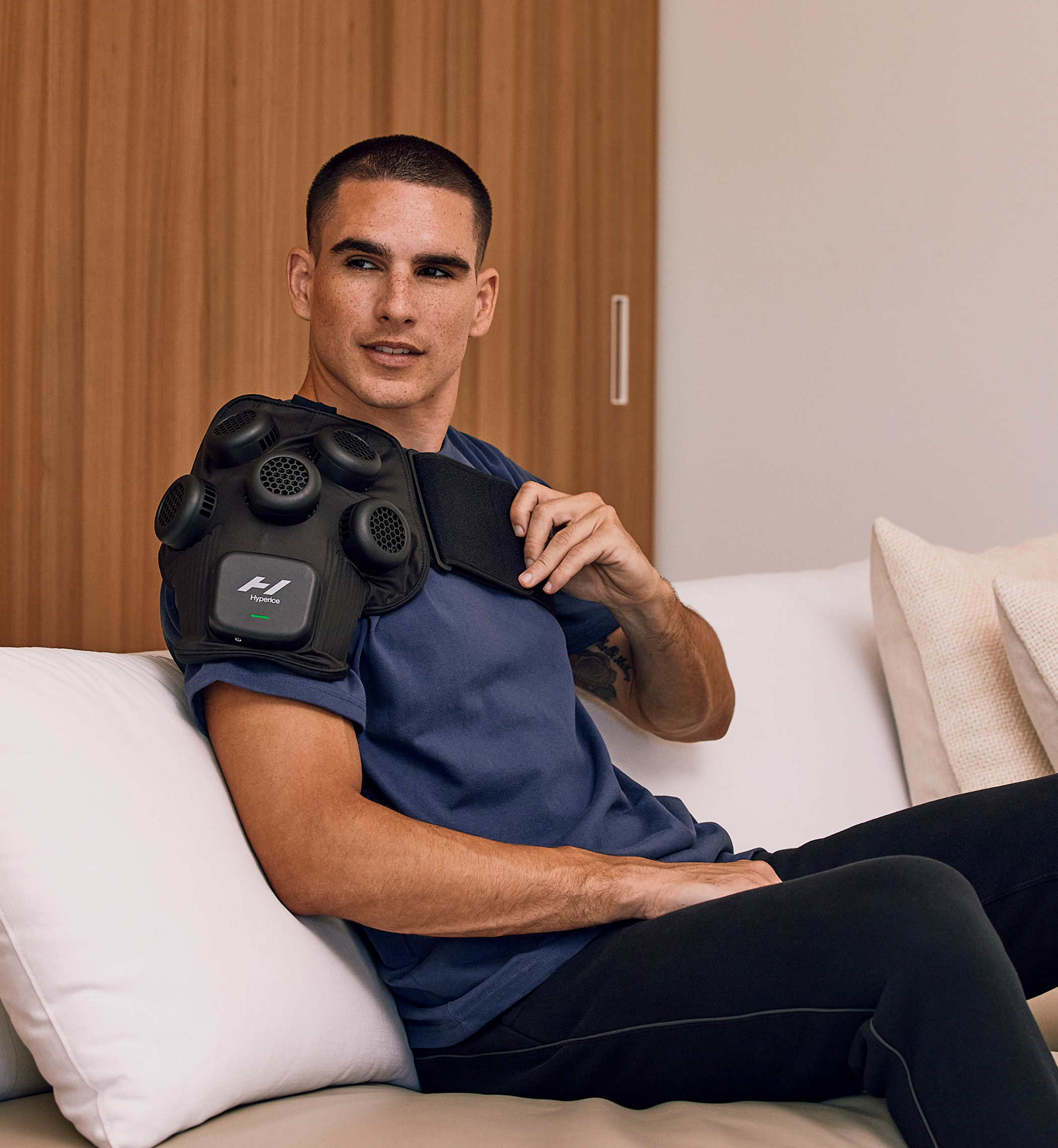 Man sitting on a couch using a black Hyperice X device on his shoulder.