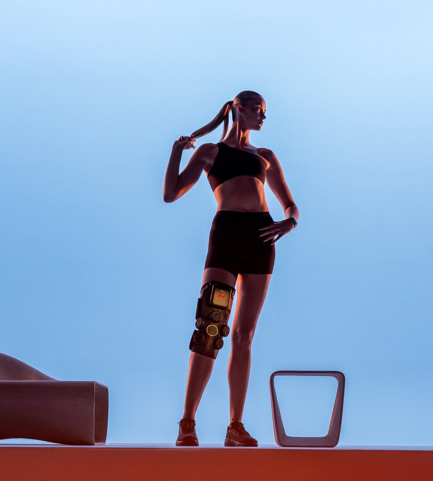 Woman in athletic wear with a Hyperice X 2 Knee device against a blue sky, like background