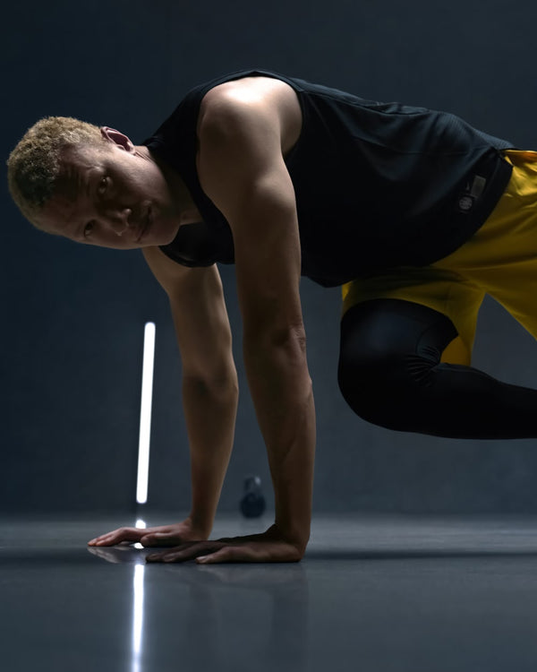 Person in a black tank top and yellow pants performing a workout in a pushup position against a dark background with a single light source.