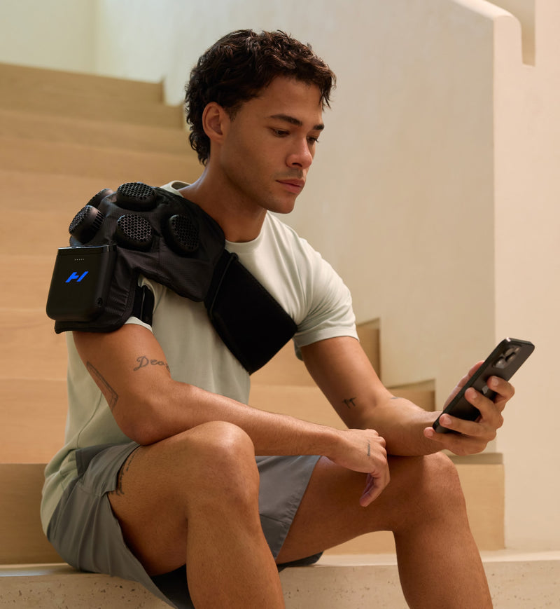 Man sitting on steps using a smartphone with a hyperice x 2 shoulder on his shoulder.