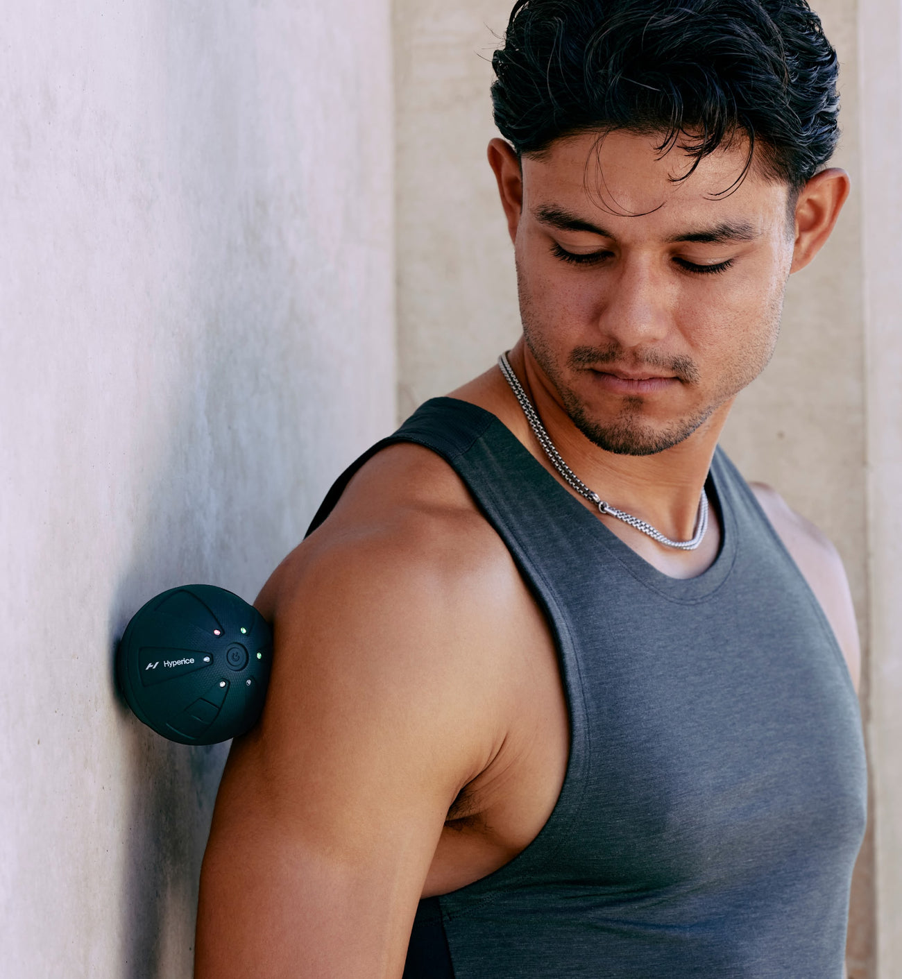 Man wearing a gray tank top pressing a hypersphere go between his shoulder and a light-colored wall.