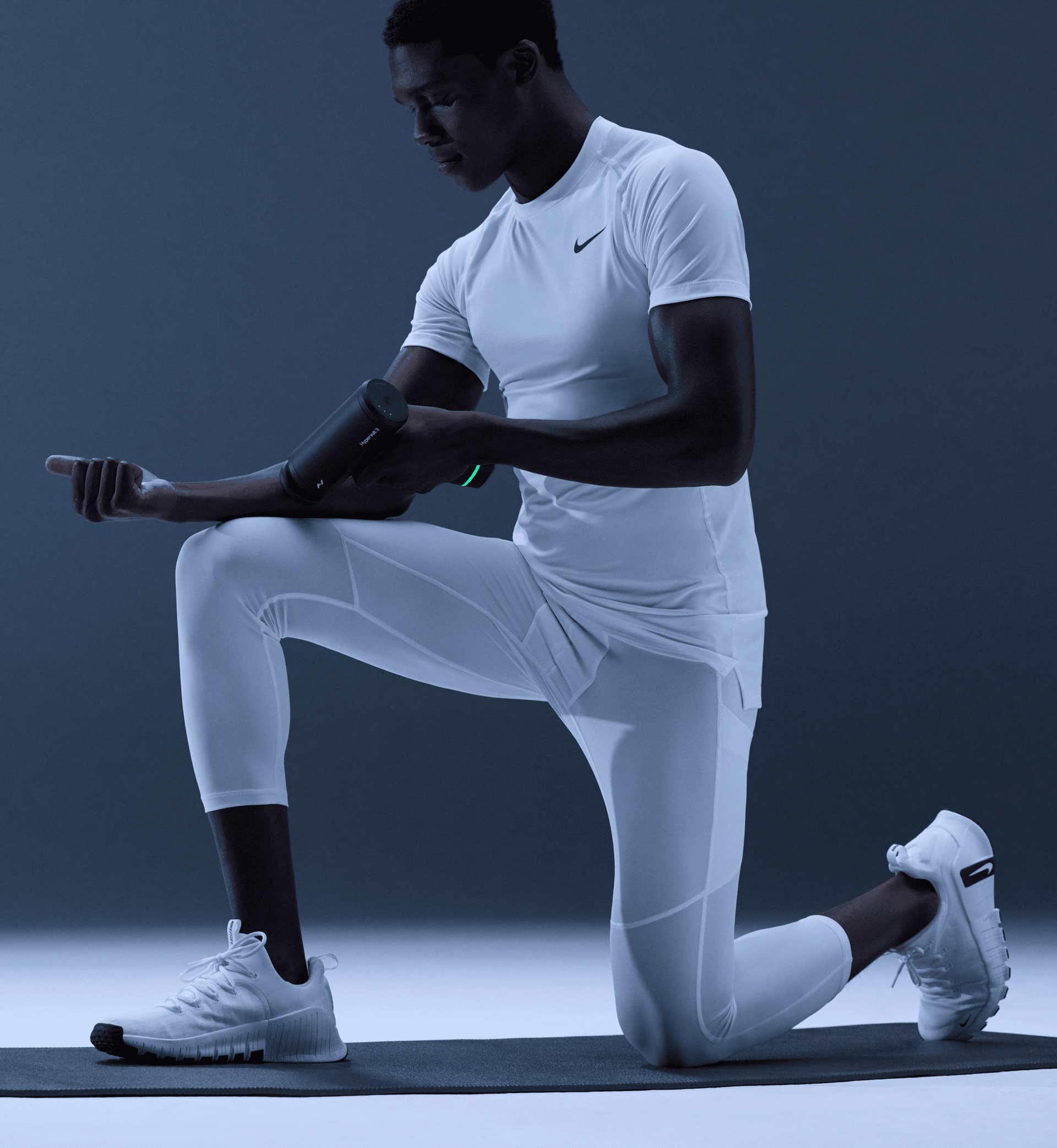Person in white athletic wear performing a lunge while resting their arm on their leg so they can use a Hypervolt 3 Massage device on their arm, all on a dark background