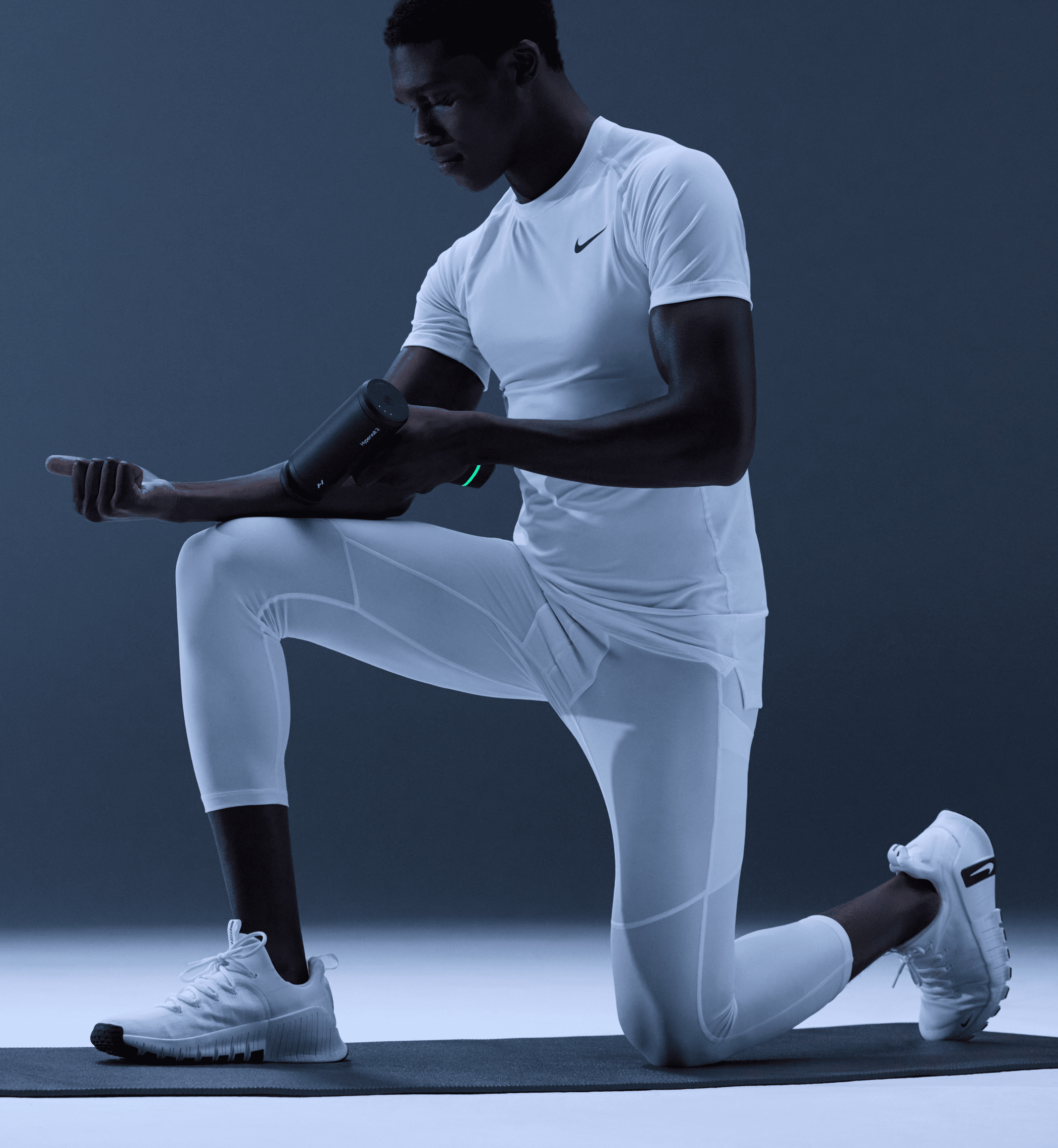 Person in white athletic wear performing a lunge while resting their arm on their leg so they can use a Hypervolt 3 Massage device on their arm, all on a dark background