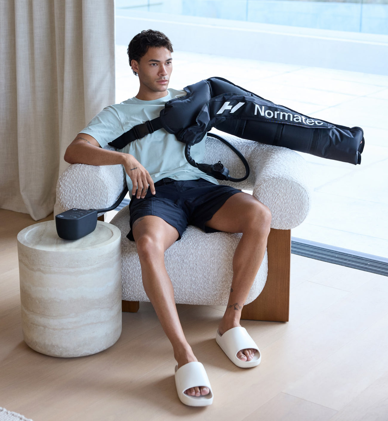 Man sitting on a chair with a Normatec 3 arm attachment on his arm, in a modern indoor setting.