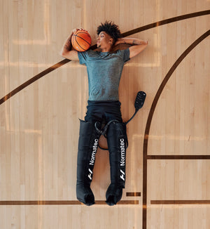 Person lying on a basketball court holding a ball, wearing Normatec 3 legs compression sleeves