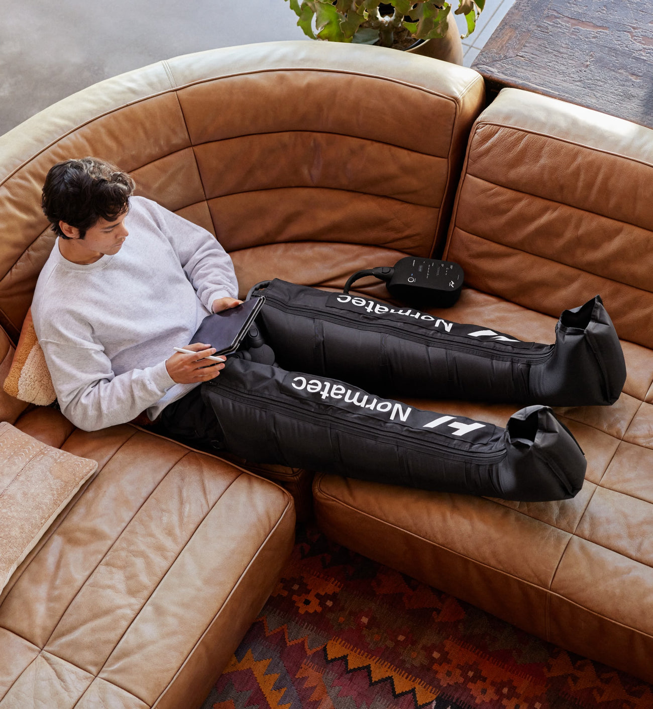 Person sitting on a brown leather sofa with a Normatec 3 legs device on their legs.