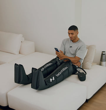 Man sitting on a white couch using a smartphone while he is wearing the Normatec 3 Dymanic air compression  leg system