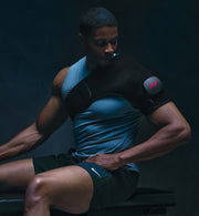 Personne portant une tenue de sport noire et bleue avec un sac à dos Venom 2, assise devant un fond sombre.